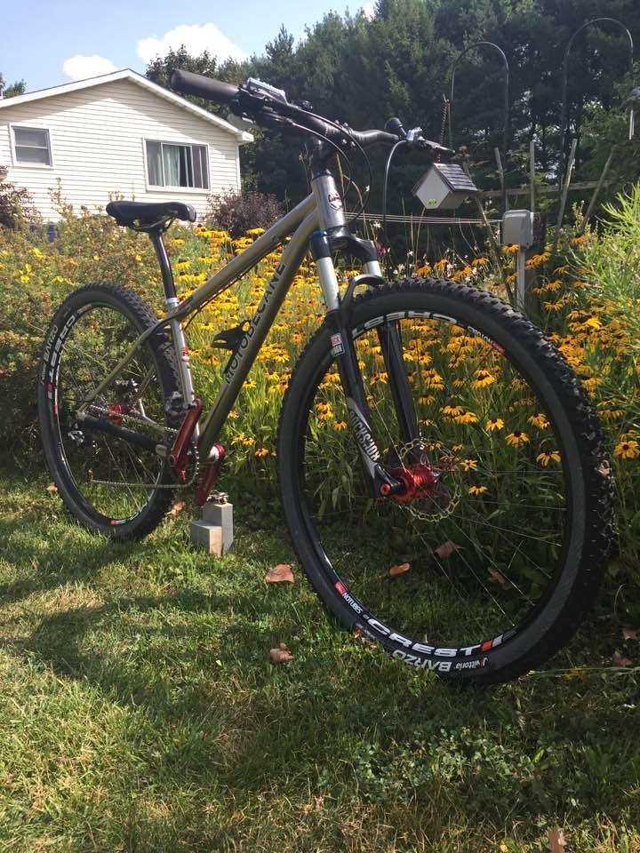 Motobecane Fly Team Ti 29er: A mountain bike with a gray and red frame is parked on green grass in front of a house, surrounded by yellow flowers. The bike features thick tires and front suspension, indicating it's designed for off-road use. The background shows a grassy area and a small garden.