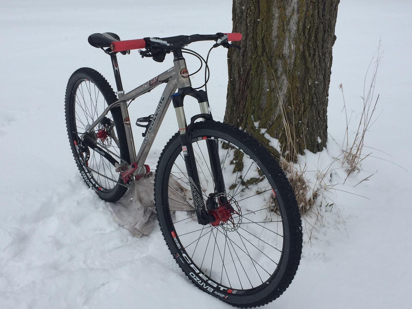 Motobecane Fly Team Ti 29er: A mountain bike resting against a tree in a snowy landscape, featuring a silver frame, red handlebars, and rugged tires designed for winter conditions. The ground is covered in fresh snow, and there are some patches of grass visible nearby.