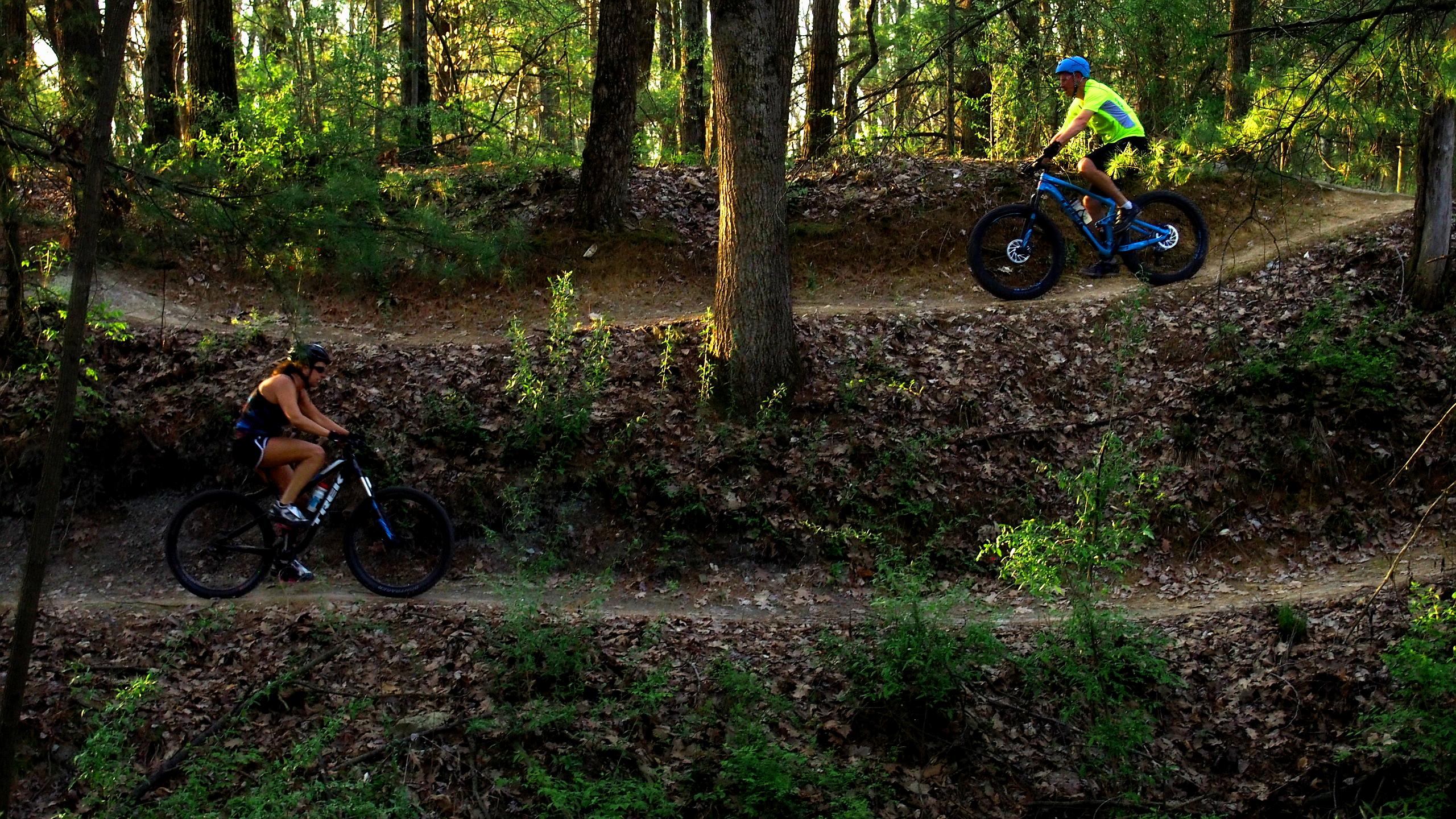 Two mountain bikers navigate a winding dirt trail through a wooded area, surrounded by tall trees and greenery. One cyclist is riding uphill on the right, wearing a bright yellow shirt and a blue helmet, while the other is biking downhill on the left, dressed in a tank top and shorts. Sunlight filters through the trees, casting dappled shadows on the ground. Griffin Bike Park mountain bike trail.