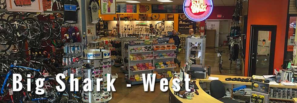 Alt text: Interior view of Big Shark - West bicycle shop, featuring a variety of bicycles, biking accessories, and merchandise displayed on shelves. A customer and staff member can be seen engaging in conversation at the checkout counter, with colorful signage and products in the background.