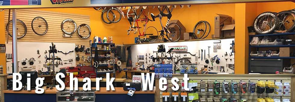 A brightly lit indoor bicycle shop featuring various bike parts, tools, and accessories displayed on racks and counters. Bicycles are hung on the wall, and shelves filled with items like bike pumps and repair kits are visible in the foreground. The store's name, "Big Shark - West," is prominently featured at the bottom of the image.