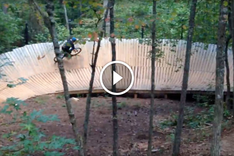 A cyclist riding along a wooden pump track in a wooded area, surrounded by trees. The track features a curved wall, allowing the rider to gain speed and perform tricks.