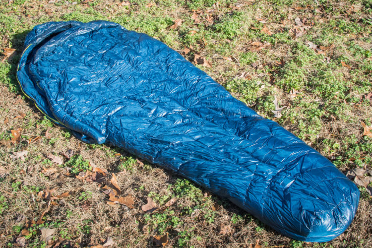 A blue sleeping bag laid out on grassy ground, surrounded by scattered leaves and patches of greenery.