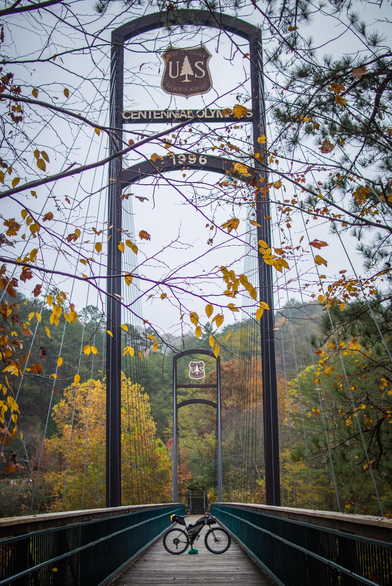 A scenic view of a suspension bridge with a sign reading "Centennial Olympics 1996" and "U.S. Forest Service." The bridge spans a wooded area with colorful autumn foliage. A bicycle is parked on the bridge, framed by bare branches. Tanasi Trail System mountain bike trail.