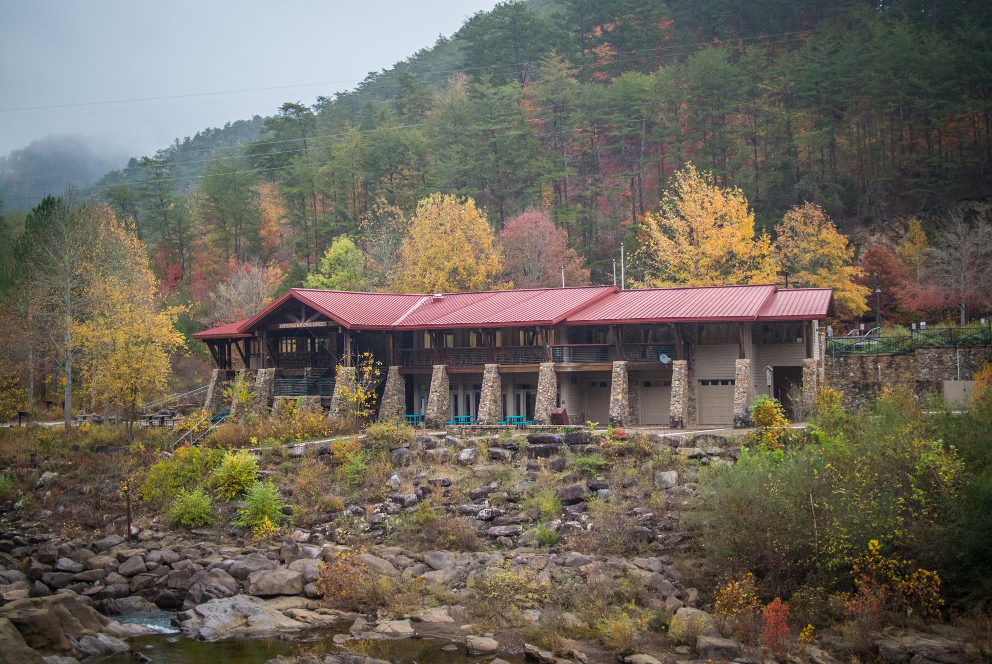 A rustic building with a red metal roof situated along a riverbank, surrounded by colorful autumn foliage and hills in the background. Stone pillars support the structure, and there are outdoor seating areas visible. The scene captures the essence of a tranquil natural environment. Tanasi Trail System mountain bike trail.
