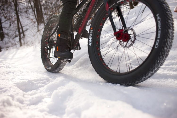 A close-up view of a fat bike riding on a snowy trail. The focus is on the rear wheel, showcasing the wide tire designed for snow, with a rider's foot visible on the pedal. Snow is kicked up around the bike as it moves through a winter landscape, with trees blurred in the background.