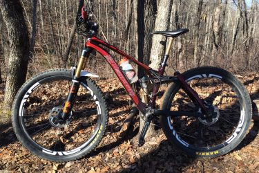 A red and black mountain bike parked on a forest trail surrounded by trees and fallen leaves. The bike features thick tires, a suspension fork, and a prominent brand logo on the frame. Sunlight filters through the trees, illuminating the scene. Bracken Preserve mountain bike trail.