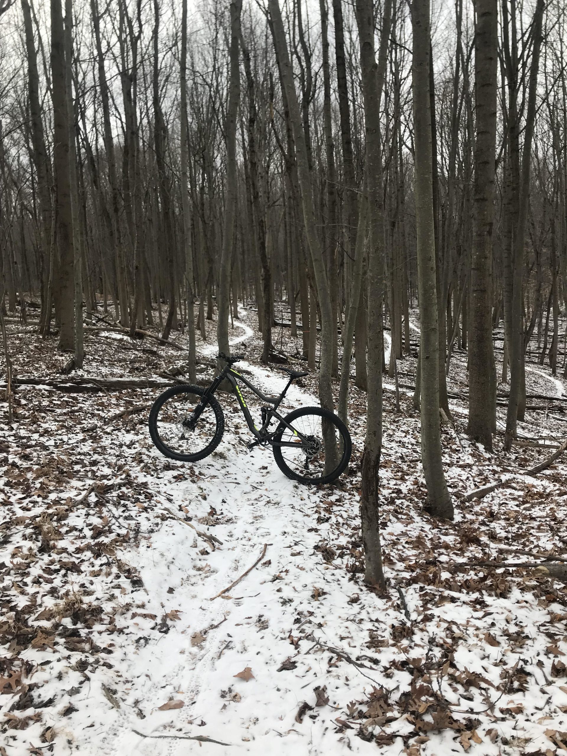 A mountain bike resting on a snow-covered trail in a wooded area, surrounded by bare trees and fallen leaves. The path winds through the forest, with snow highlighting the ground and adding to the serene winter atmosphere. Freedom Center mountain bike trail.