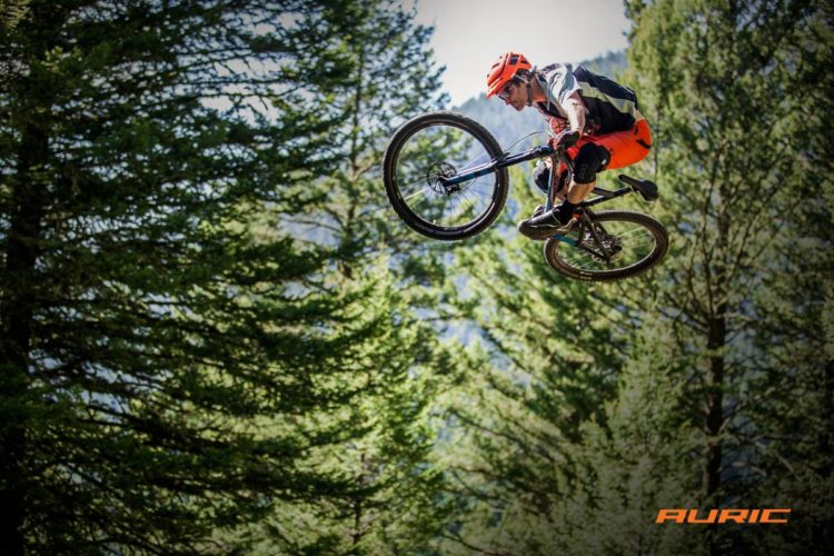 A mountain biker performs an airborne trick while jumping off a ramp in a forested area. The rider is wearing bright orange gear, including a helmet, and is captured mid-air with the bike's front wheel lifted. Tall evergreen trees create a natural backdrop, highlighting the outdoor adventure.