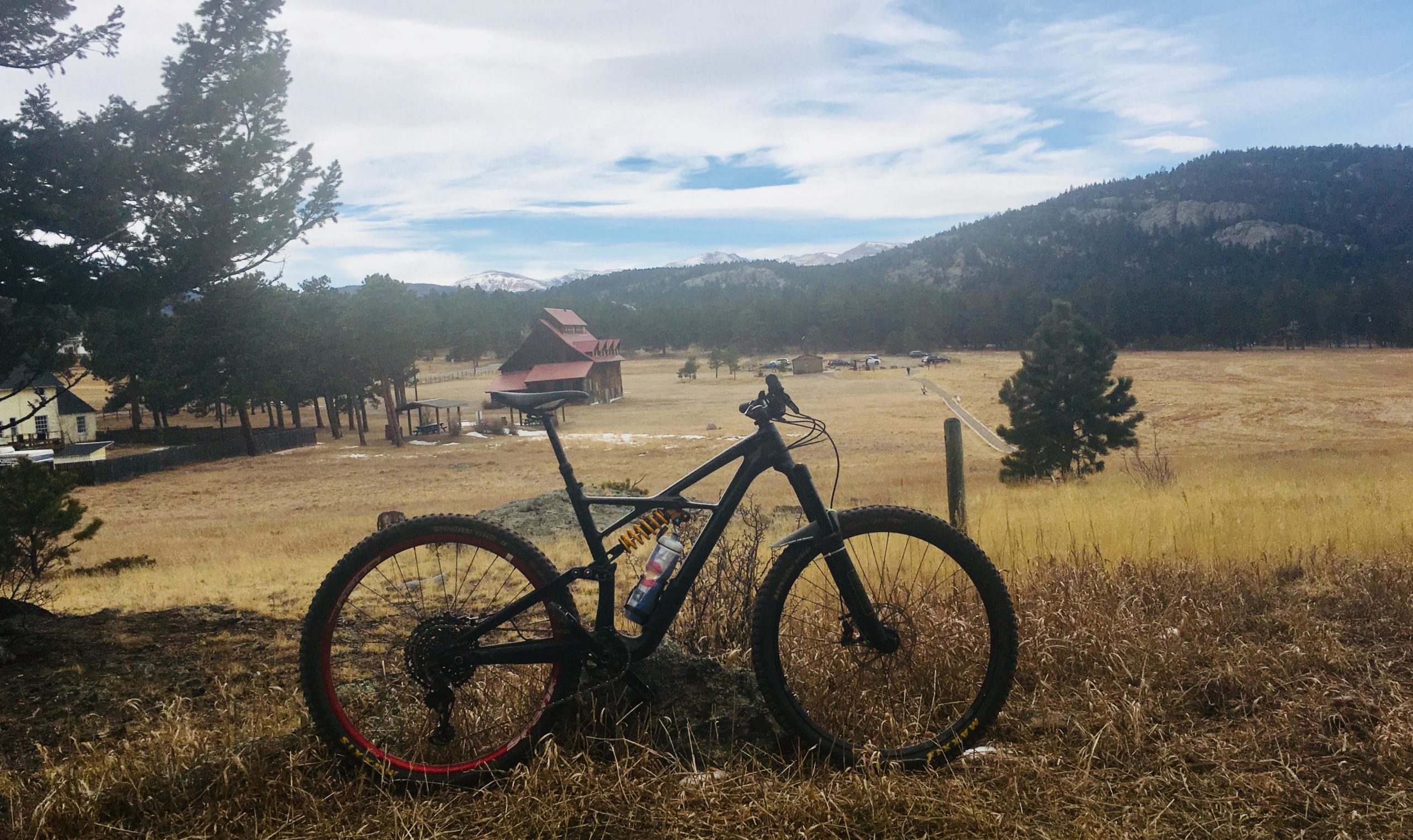 A mountain bike rests on a grassy patch overlooking a scenic landscape featuring a fenced pasture with a red barn, surrounded by trees and distant mountains under a cloudy sky. 3 Sisters / Alderfer mountain bike trail.