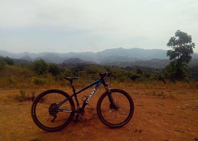 Giant ATX 27.5 2: A mountain bike parked on a dirt path with a scenic view of mountainous terrain in the background. The landscape features rolling hills and sparse vegetation under a cloudy sky.