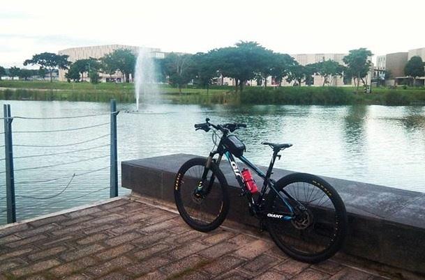 Giant ATX 27.5 2: A black mountain bike resting against a low stone wall by a calm lake, with a fountain in the background. Lush greenery surrounds the water, and a building is visible in the distance under a cloudy sky.