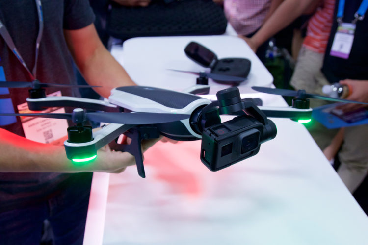 A person holding a drone with a camera attached, showcasing its features at a technology exhibition. The drone has green LED lights and is positioned above a table with various devices in the background, with attendees visible.