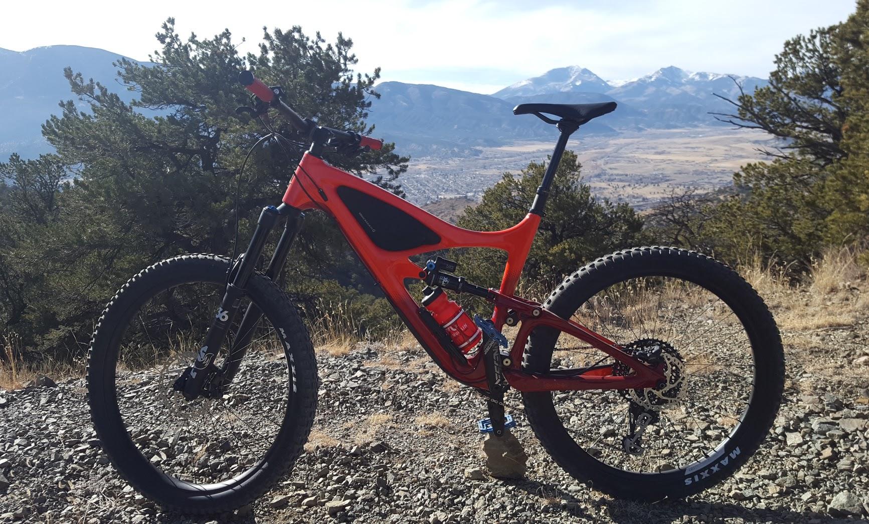 A bright orange mountain bike is positioned on a rocky trail with a scenic view of mountains and a valley in the background. The bike features wide tires, front suspension, and a water bottle mounted on the frame, surrounded by greenery and dry grass. Cottonwood mountain bike trail.