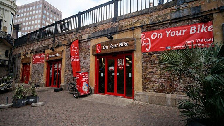 Image of the storefront for "On Your Bike," featuring red banners with the shop name. The brick wall building has large glass-paneled doors and windows, with outdoor planters and a bicycle displayed outside. The area is paved, indicating a commercial setting.