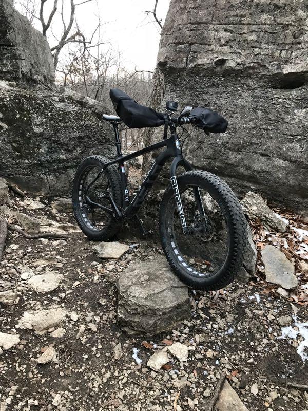 A black fat tire bike with mounted bags stands on a rocky, uneven trail surrounded by large boulders and trees. The ground is covered with scattered rocks and dried leaves, indicating a rugged outdoor environment. Swope Park Trail mountain bike trail.