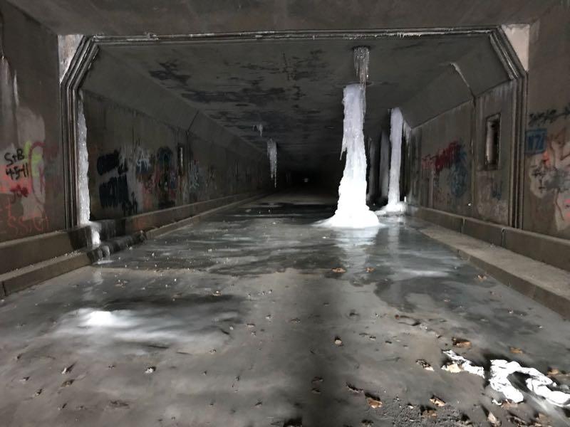 A dimly lit underpass featuring a frozen surface with patches of ice, icicles hanging from the ceiling, and walls adorned with colorful graffiti. Fallen leaves are scattered on the ground, creating a contrast with the icy scene. Pike 2 Bike mountain bike trail.