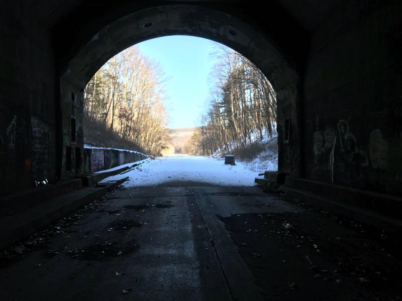 A view from inside a dark, weathered tunnel, looking out onto a snow-covered road surrounded by barren trees. The ground shows signs of autumn leaves, and faint graffiti can be seen on the tunnel walls. Sunlight streams through the tunnel opening, illuminating the path ahead. Pike 2 Bike mountain bike trail.