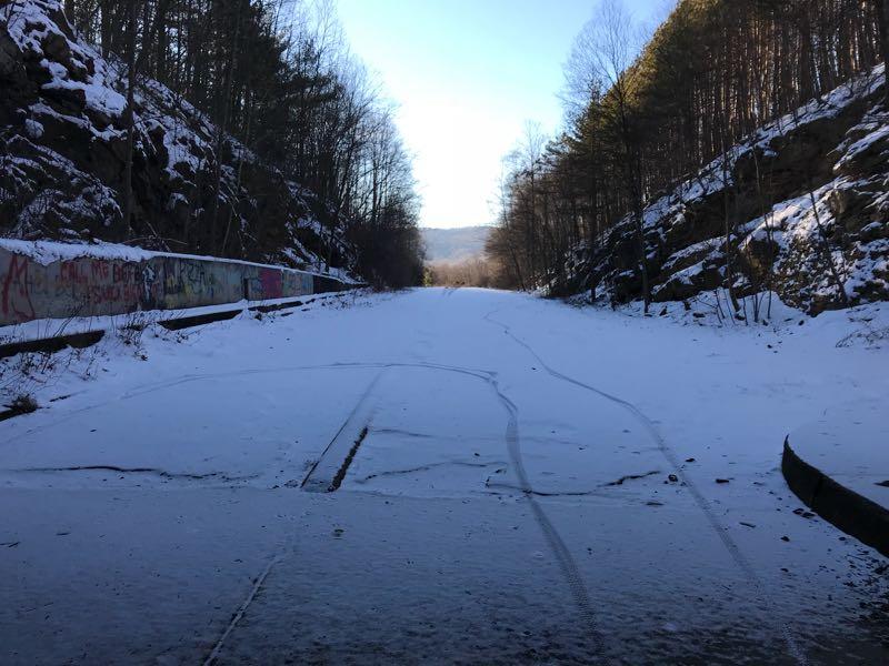 A snow-covered, abandoned railway track runs through a forested area, flanked by rocky cliffs. The scene is illuminated by bright sunlight, casting shadows on the snow. Graffiti is visible on the side of the track, and a clear blue sky is visible in the background. Pike 2 Bike mountain bike trail.