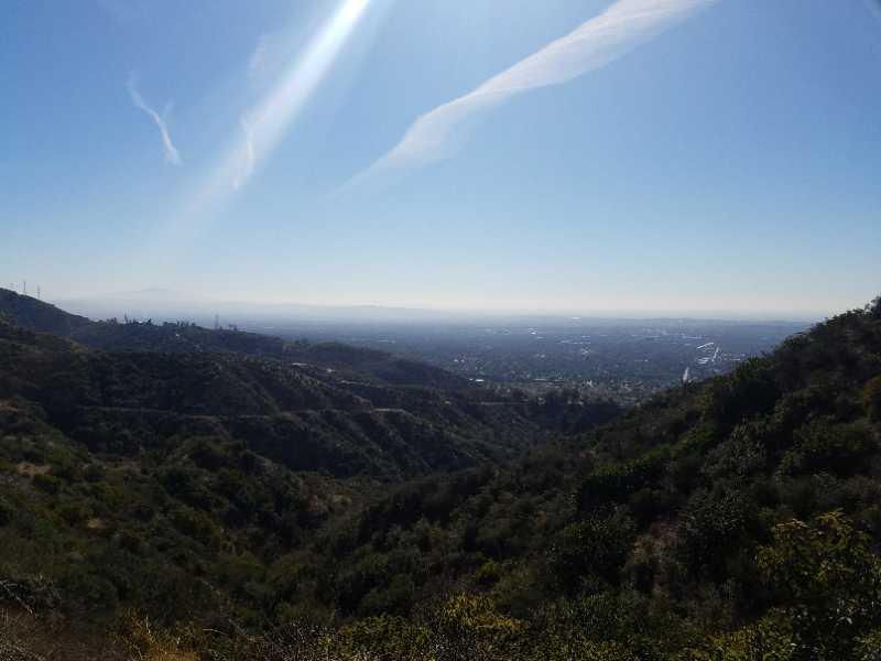 A panoramic view of a lush, green valley with rolling hills under a clear blue sky. In the distance, a hazy urban landscape can be seen, with faint outlines of buildings against the horizon. The scene captures the natural beauty of the landscape, blending vegetation with a glimpse of the city below. El Prieto mountain bike trail.