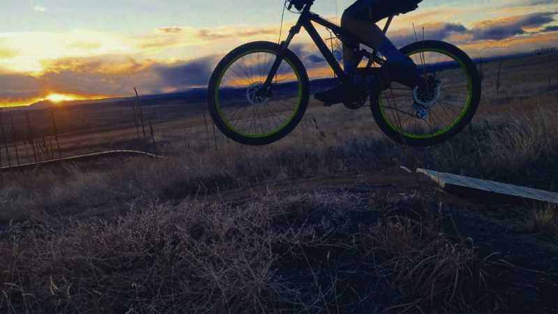 A silhouetted mountain biker performing a jump over a wooden ramp against a vibrant sunset backdrop, with grassy terrain stretching out in the foreground and a cloudy sky illuminating the scene. Bayou Gulch Bike Park mountain bike trail.
