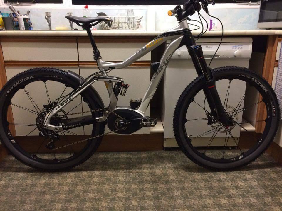 Haibike SDURO AllMtn Pro: A modern mountain bike with a sleek silver and black frame, thick tires, and advanced components, positioned indoors against a backdrop of kitchen appliances. The bike features front suspension, a visible motor, and a chain designed for off-road riding.