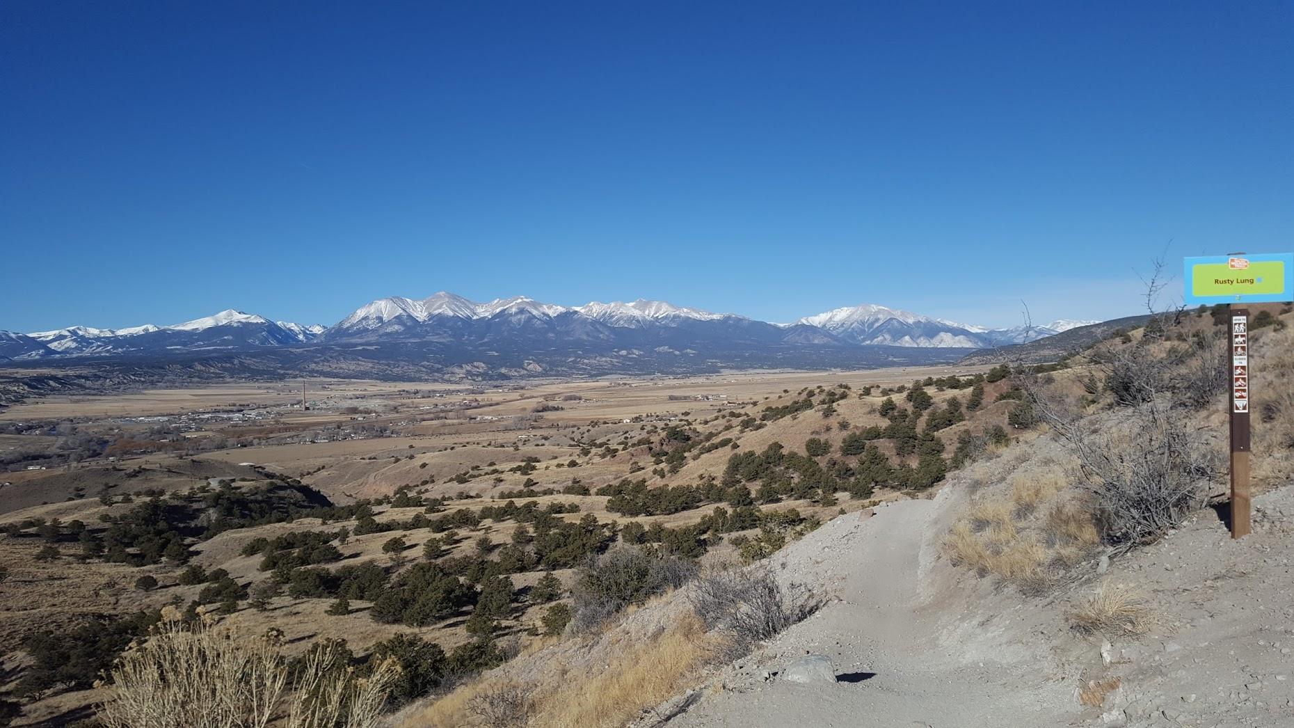 Scenic view of snow-capped mountains under a clear blue sky, overlooking a valley with sparse foliage and a trail sign labeled "Rusty Lung" on the right side. The landscape features rolling hills and a distant town, highlighting the natural beauty of the area. Rusty Lung mountain bike trail.
