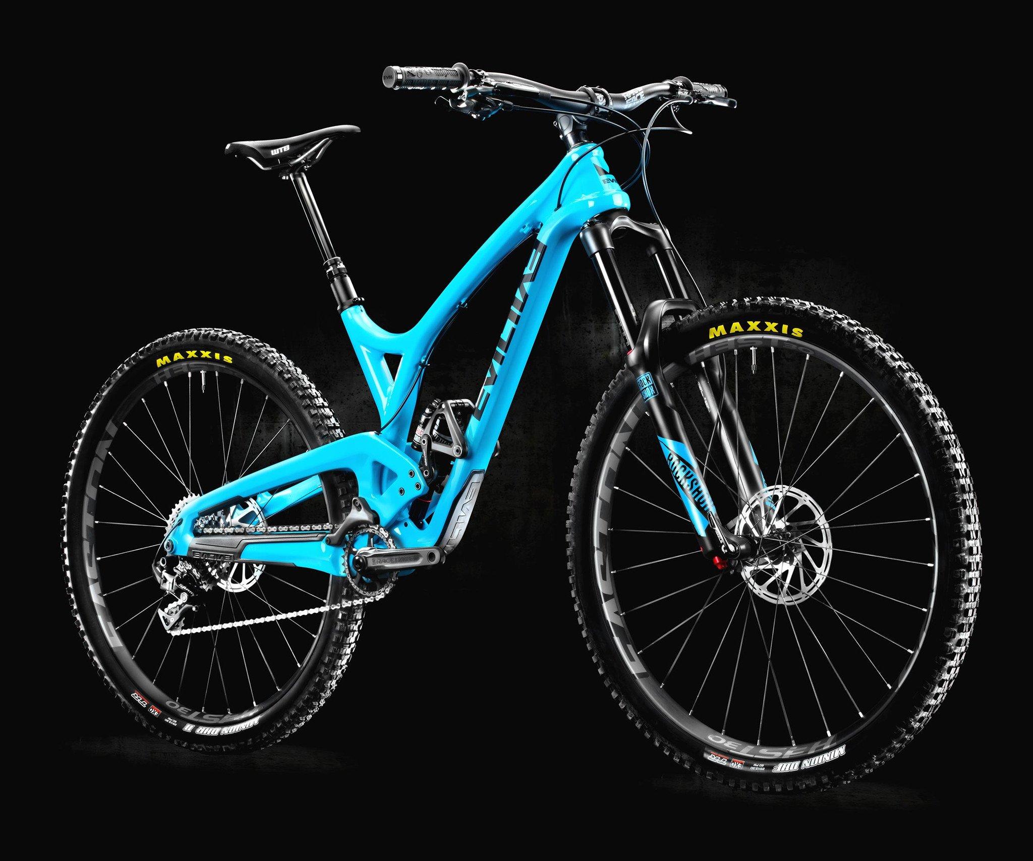 Evil Wreckoning: A bright blue mountain bike with a sleek frame and black components is showcased against a dark background. The bike features a front suspension fork, thick tires with "Maxxis" branding, and a modern design, suitable for off-road riding. The bike's saddle is positioned at a high setting, indicating readiness for action.