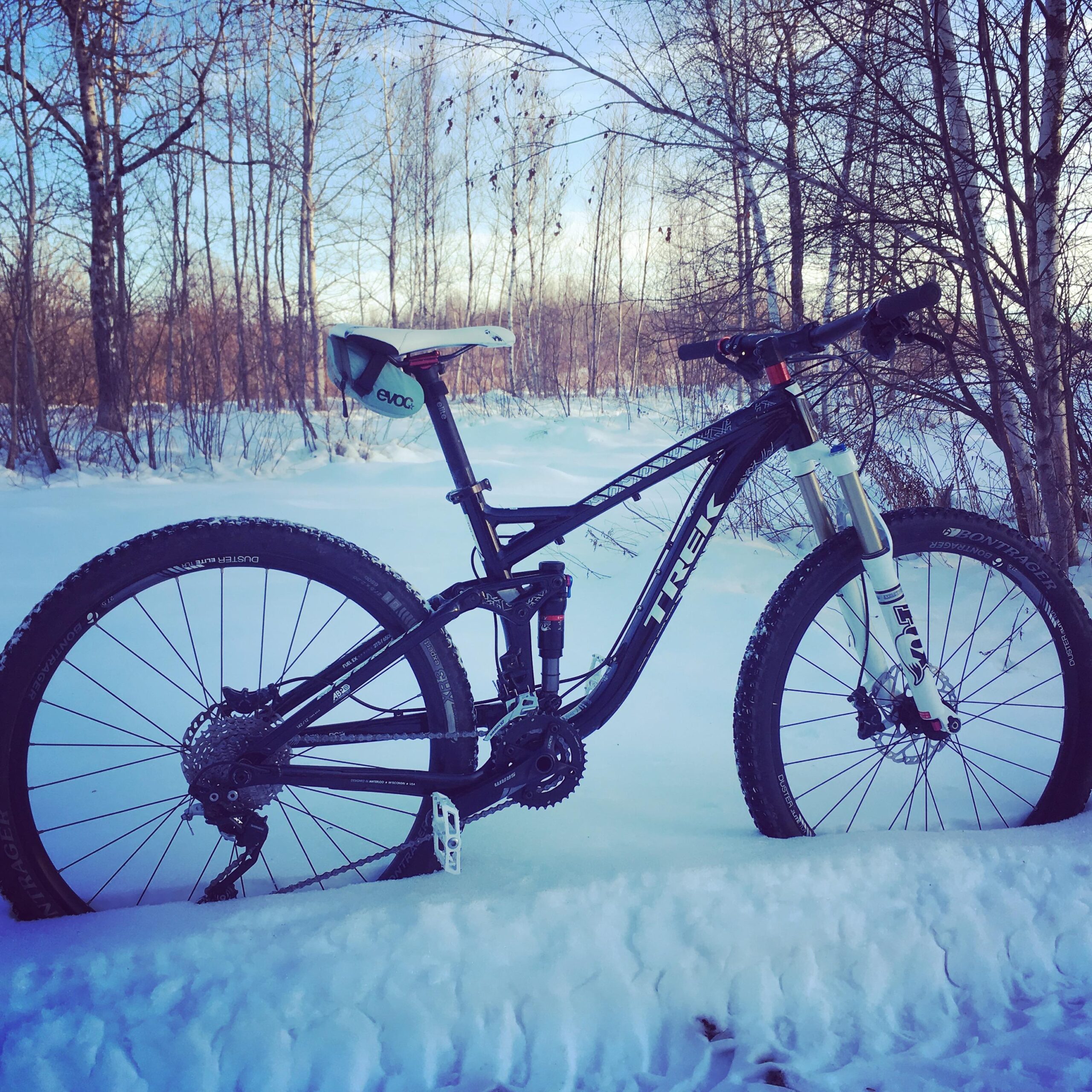 Trek Fuel EX 8: A mountain bike leaning against a snow-covered landscape, surrounded by bare trees under a clear sky. The bike features a black frame with the brand name visible, and snow covers the ground partly obscuring its wheels.