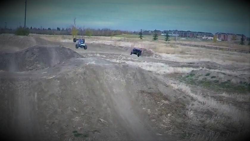 Two remote-controlled cars are airborne as they jump over dirt mounds at an outdoor track. One car is performing a flip, while the other is in mid-air, showcasing high-speed action in a grassy, open area with a cloudy sky in the background. devin mountain bike trail.