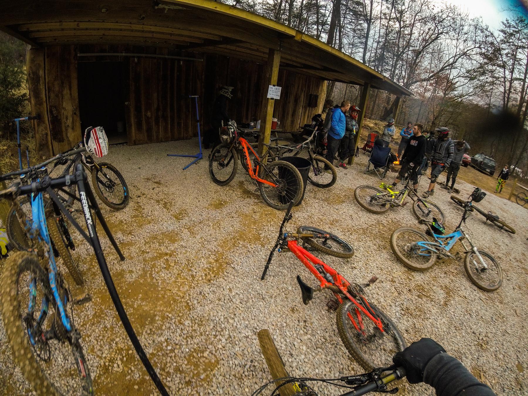 A group of mountain bikers gathered outside a rustic wooden building, with several bicycles parked on the ground around them. The scene shows a mix of bike colors, including blue, orange, and black, set against a gravel surface. Bikers in casual clothing are engaged in conversation, while bicycle repair stands are visible nearby. The surrounding area is wooded, hinting at a natural outdoor environment. Windrock Bike Park mountain bike trail.