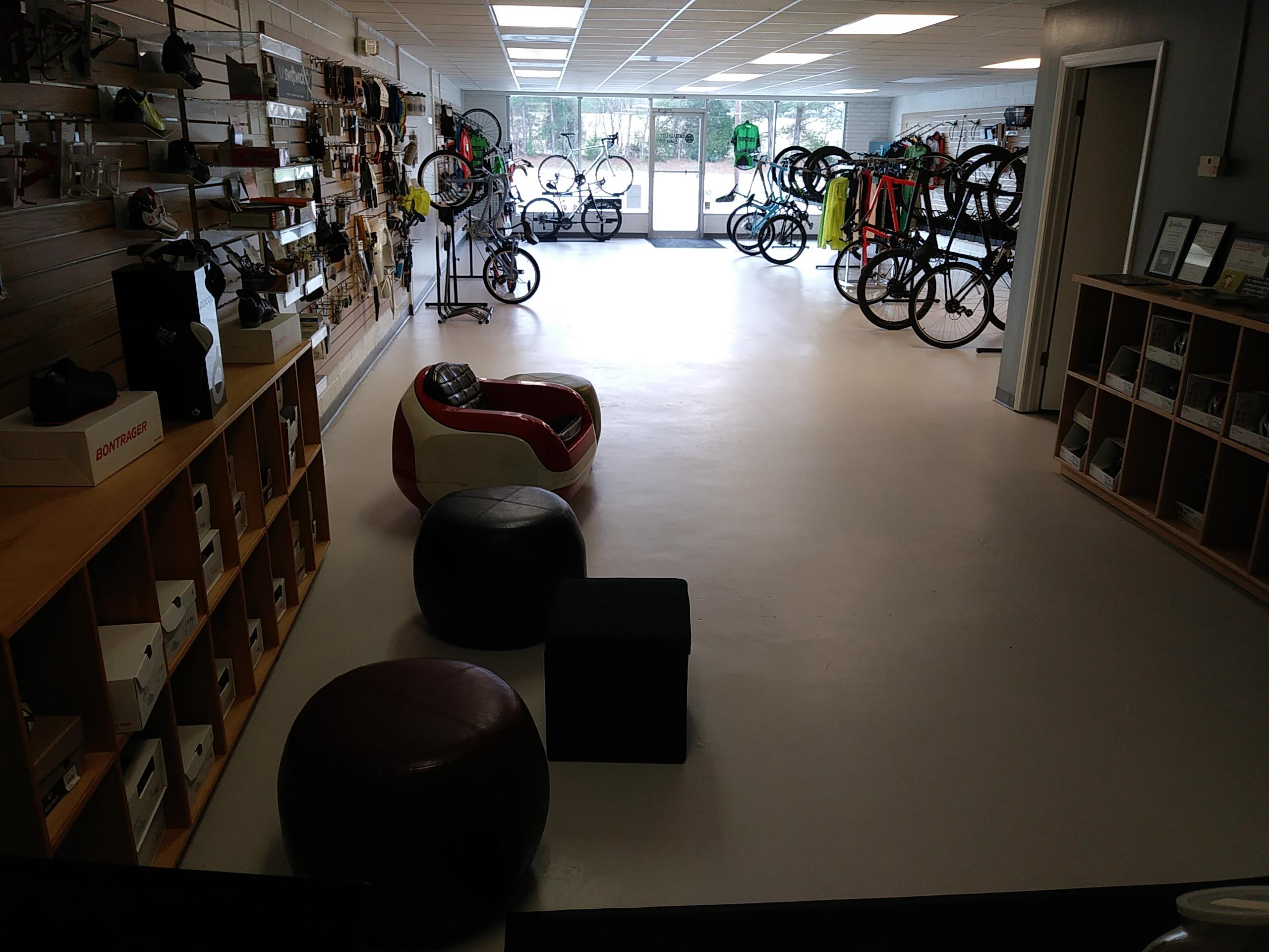 A view of a bicycle shop interior featuring a variety of bicycles displayed on racks against the wall. There are shelves on the side showcasing cycling accessories, shoes, and equipment. In the foreground, there are seating options including a red and white chair and two round stools. Bright lighting illuminates the space, and large windows provide a view of the outside.