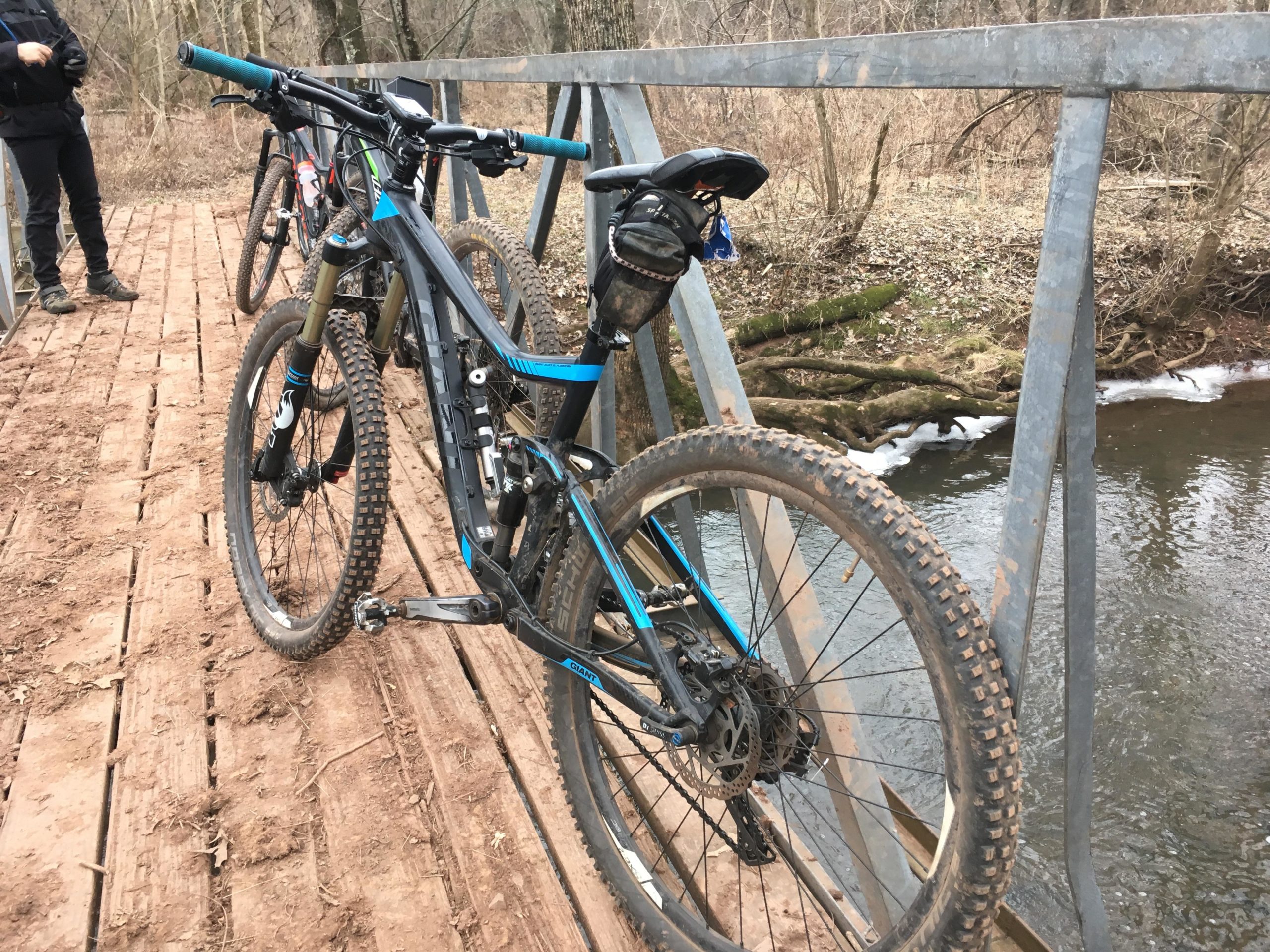 A mountain bike is secured on a wooden bridge with a creek visible below. The bike is equipped with thick tires and features a blue and black color scheme. A person in the background stands on the bridge, dressed in black, while trees and bare foliage surround the area, indicating an early spring or late winter setting. Six Mile Run mountain bike trail.