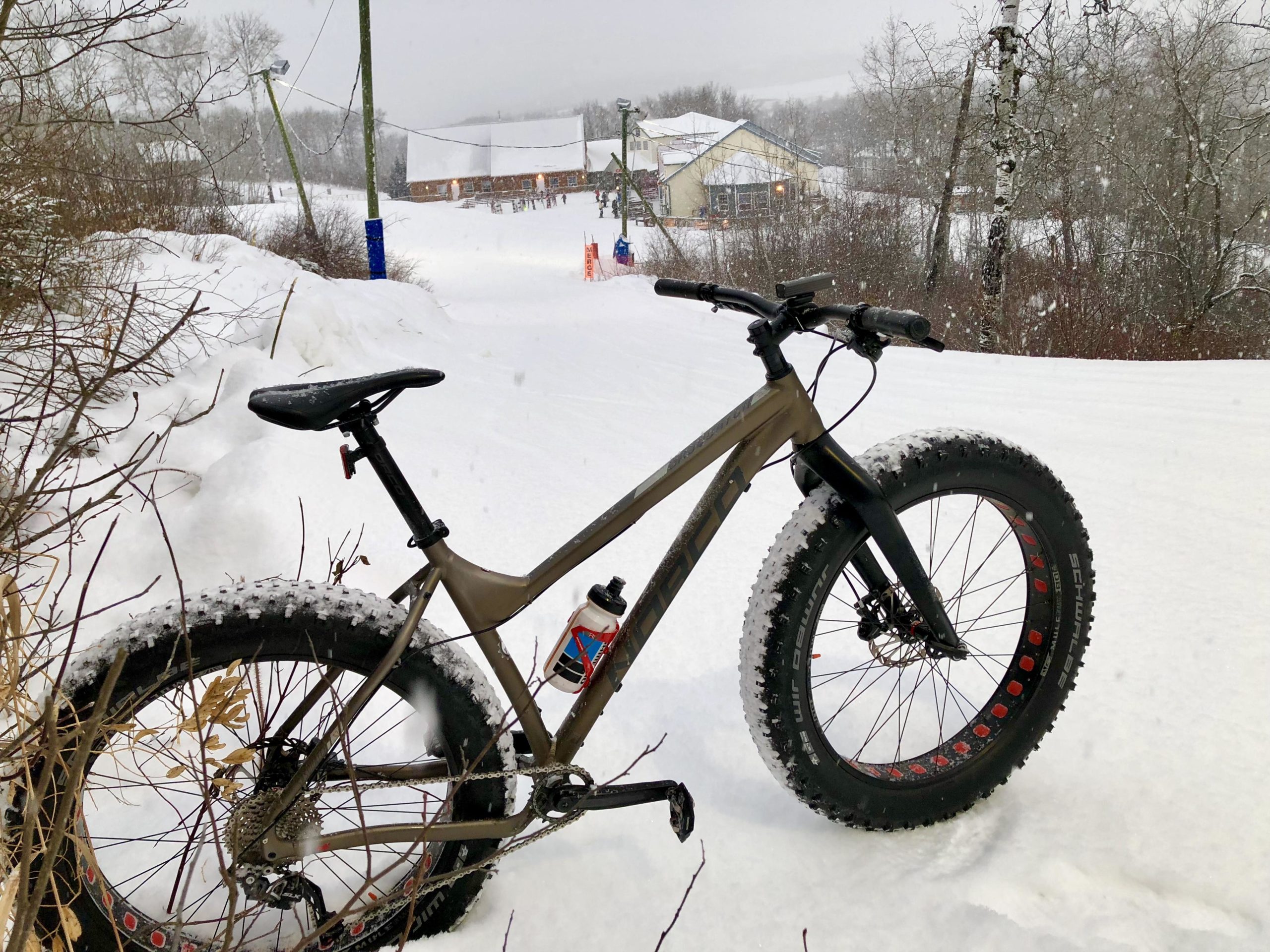 A fat bike is parked on a snowy trail, with snowflakes falling in the background. The scene depicts a winter landscape with trees and a building in the distance, suggesting a ski area or winter recreation spot. Ridge line & Home Run mountain bike trail.