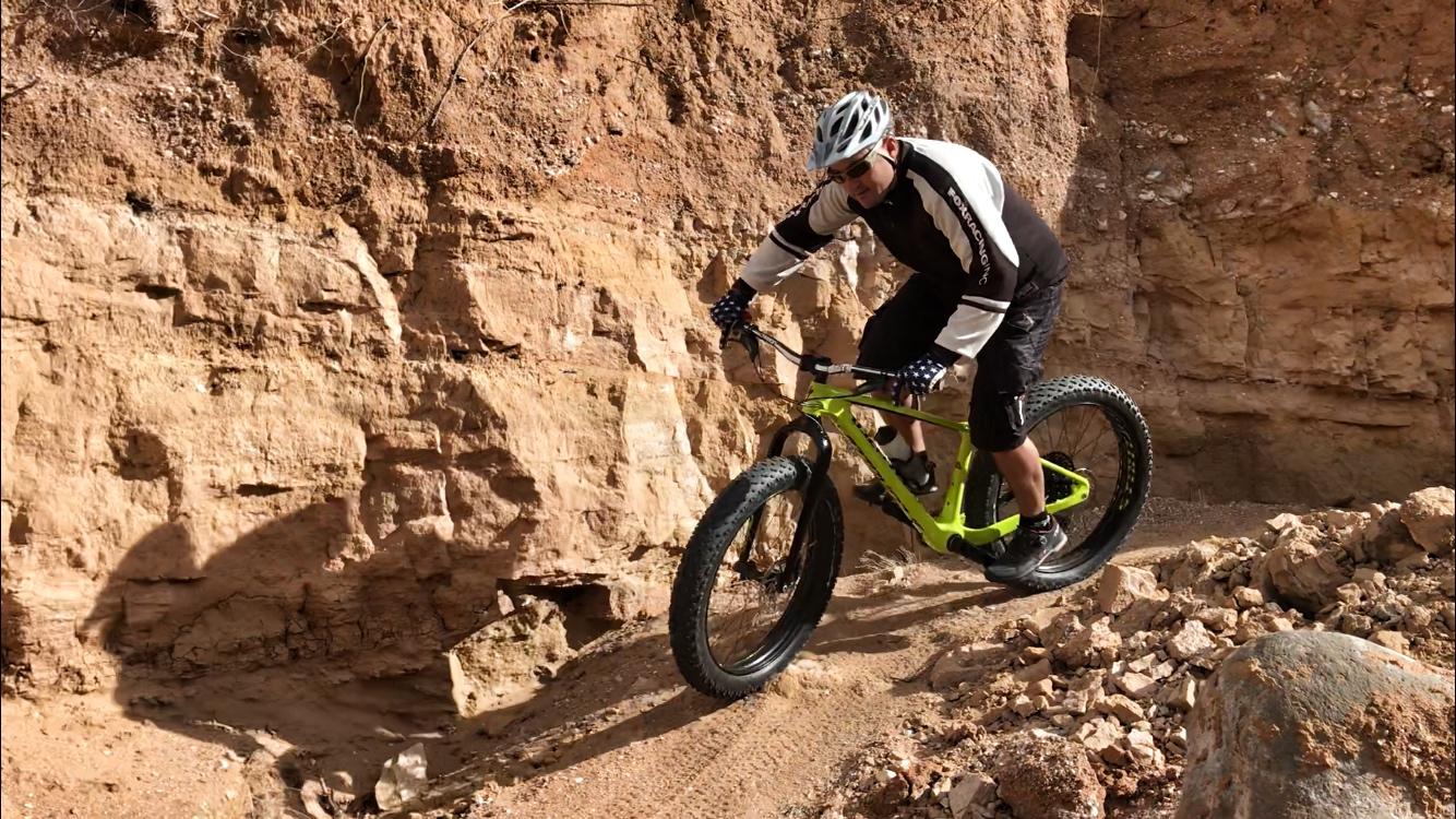 A mountain biker riding a bright green fat tire bicycle on a rocky, uneven trail surrounded by steep earthy cliffs. The cyclist is wearing a helmet and protective gear, focused on navigating the challenging terrain. Sunlight illuminates the scene, highlighting the texture of the rocks and dirt. Mariposa Fat Bike Trails mountain bike trail.