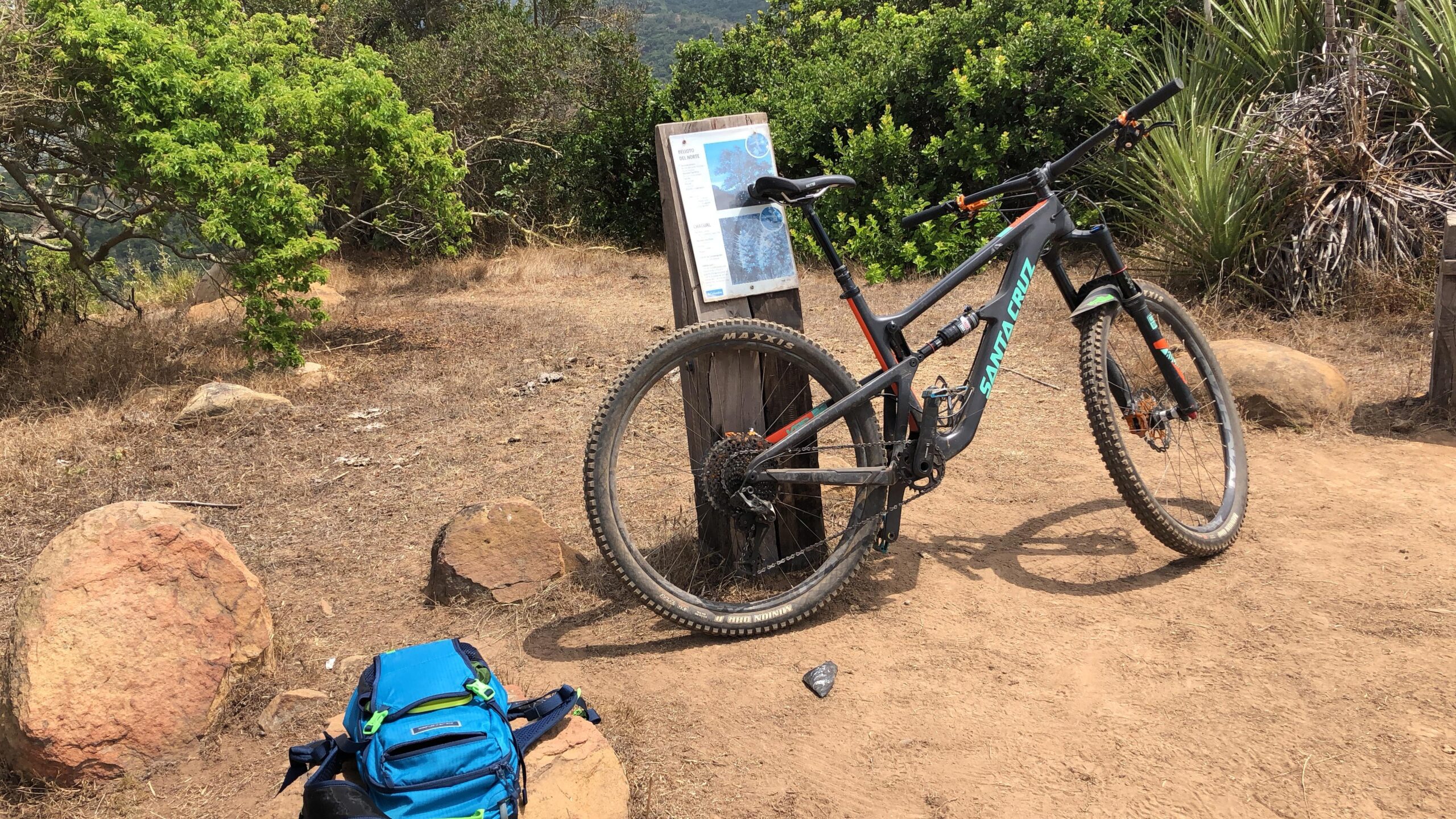 Santa Cruz Hightower 29er/27.5: A mountain bike leaning against a wooden post with an information sign, surrounded by rocks and greenery on a dirt trail. A blue backpack is placed on the ground nearby. The scene is set in a sunny outdoor environment.