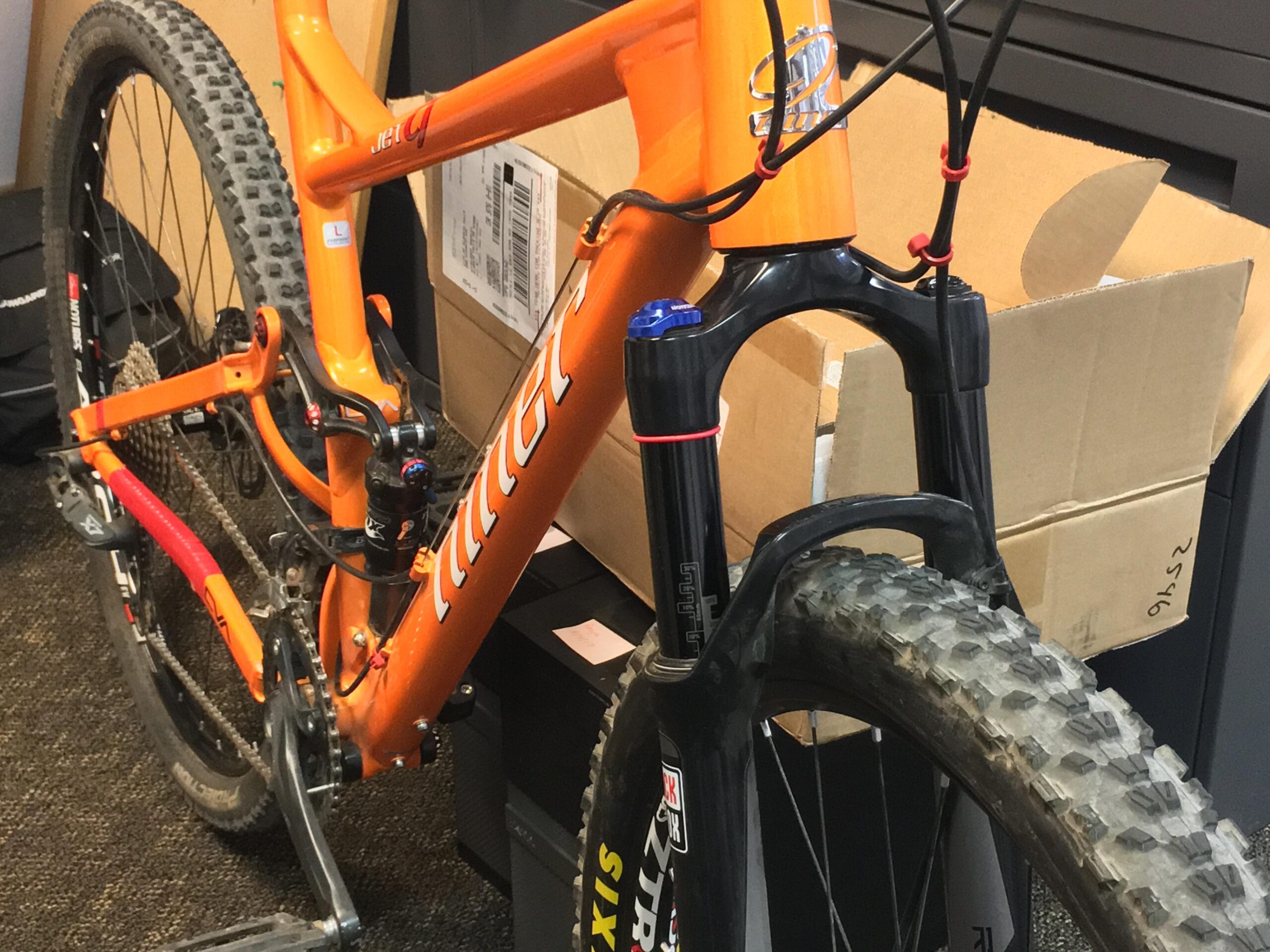 Niner Jet 9: A close-up view of an orange mountain bike frame, featuring a front suspension fork and knobby tires, set against a background of cardboard boxes. The image highlights the bike's components, including handlebars and chain, with some dirt visible on the tires.