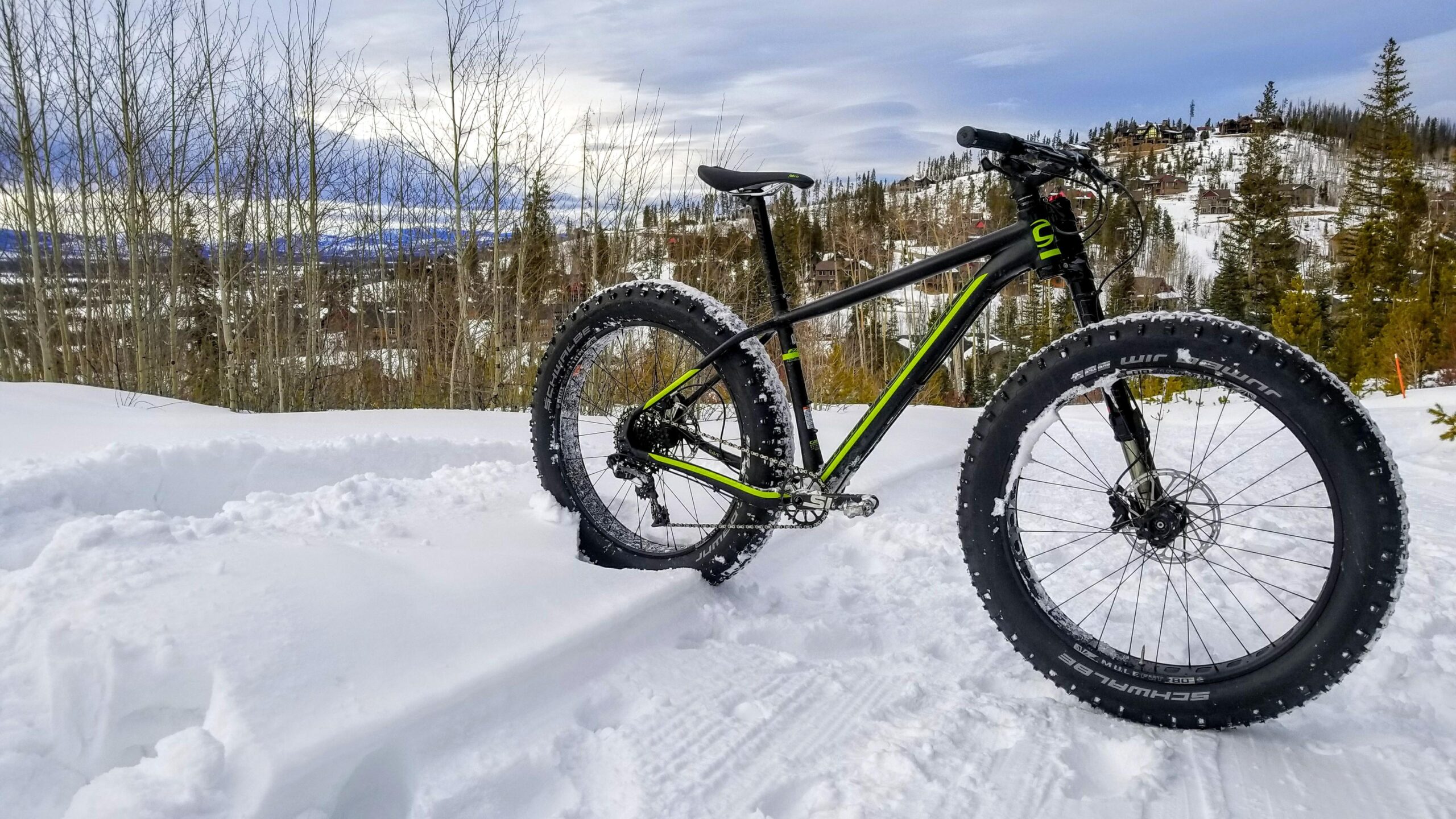 Cannondale Fat CAAD 1: Alt text: A fat tire bicycle stands on a snowy trail, surrounded by trees and a winter landscape. The bike has wide tires designed for snow, with tread patterns clearly visible, and features a black frame with bright green accents. In the background, a scenic view of hills and houses is visible under a cloudy sky.