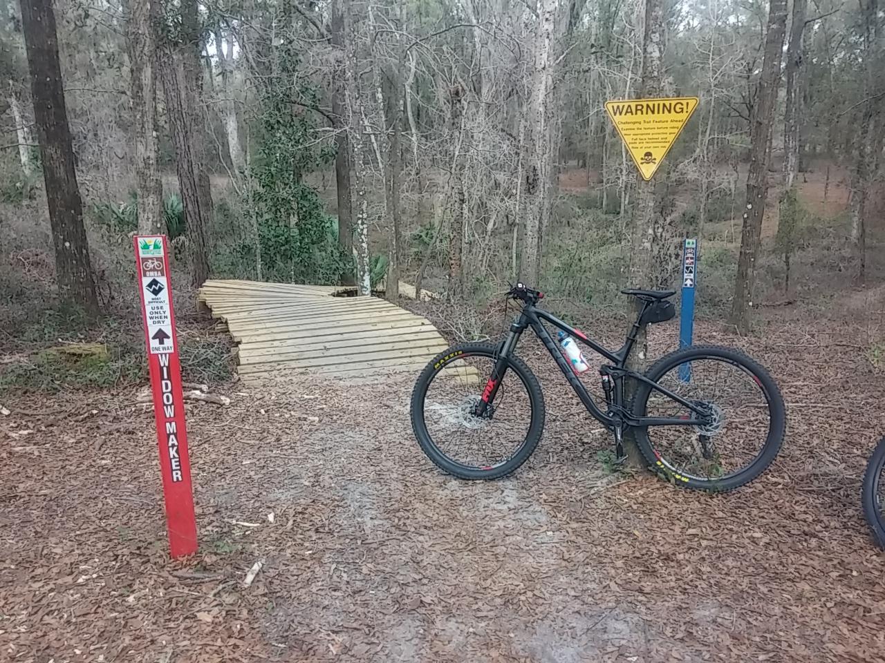 Trek EX 8.29: A mountain bike parked beside a wooden bridge on a forested trail. Trail markers indicate the path name "Widow Maker" with a warning sign for challenging trail features ahead. The surrounding area features tall trees and fallen leaves on the ground.
