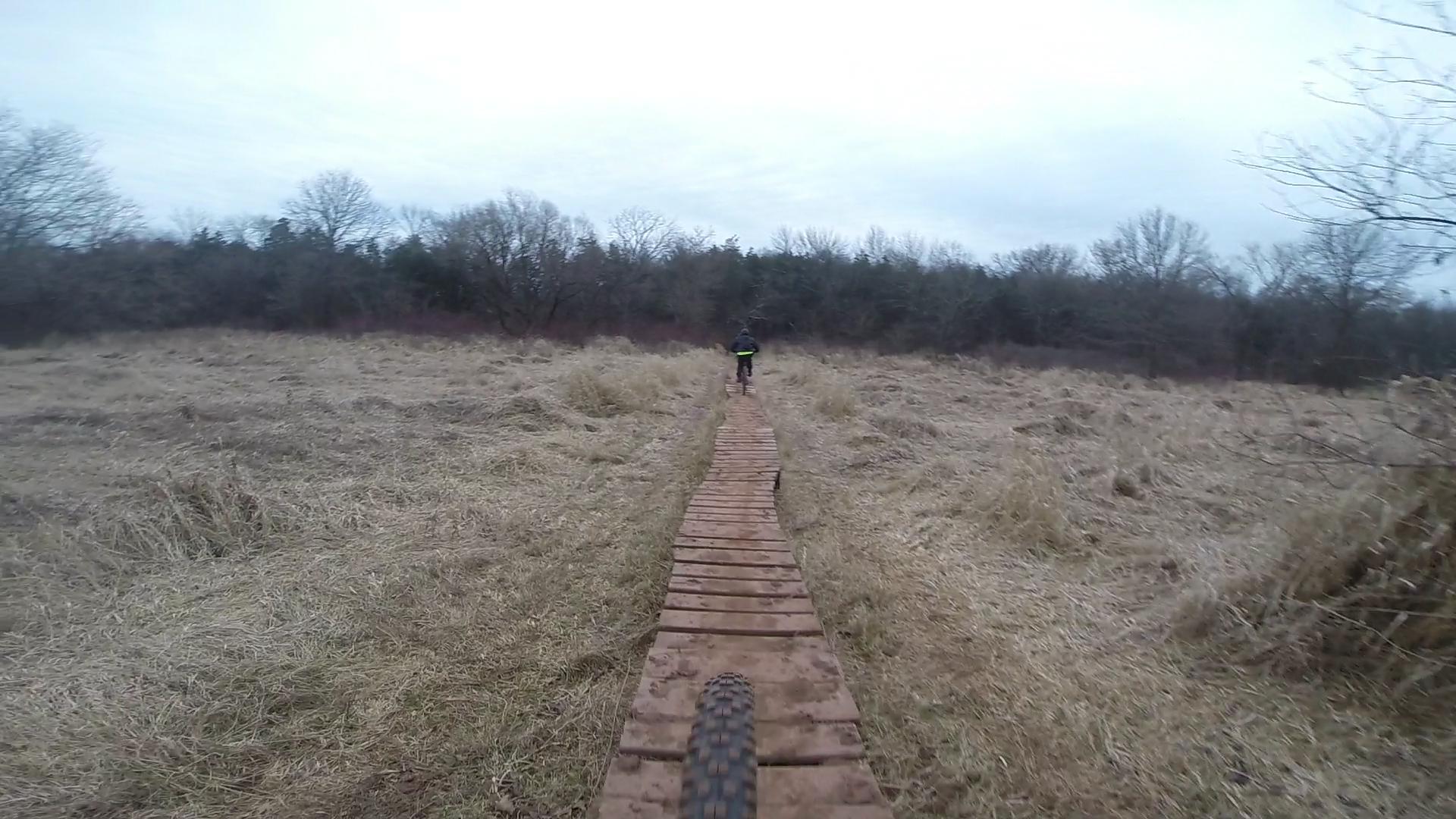 A mountain biker rides along a narrow wooden path through a grassy area, surrounded by trees under a cloudy sky. The view is from the perspective of the bike, focusing on the trail and the distant rider. Six Mile Run mountain bike trail.