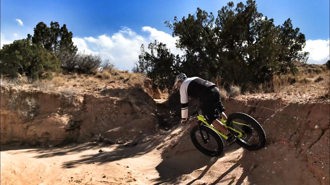 A person riding a mountain bike on a sandy trail, navigating a steep incline in a rugged outdoor environment. The cyclist is wearing a helmet and a black and white outfit, with a vivid yellow bike. The backdrop features trees and a bright blue sky with wispy clouds. Mariposa Fat Bike Trails mountain bike trail.