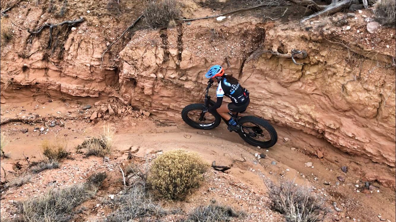 A mountain biker navigates a narrow dirt trail surrounded by steep, reddish-brown canyon walls. The biker is wearing a helmet and cycling gear, and they are riding a bike with wide tires designed for rugged terrain. Sparse vegetation, including small bushes and grass, can be seen in the arid landscape. Mariposa Fat Bike Trails mountain bike trail.