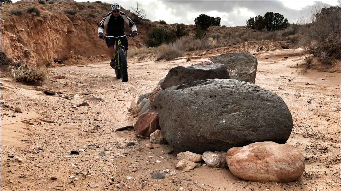 A person riding a fat bike on a sandy trail, navigating around large rocks in the foreground. The background features a rugged landscape with dry vegetation and a cloudy sky. Mariposa Fat Bike Trails mountain bike trail.