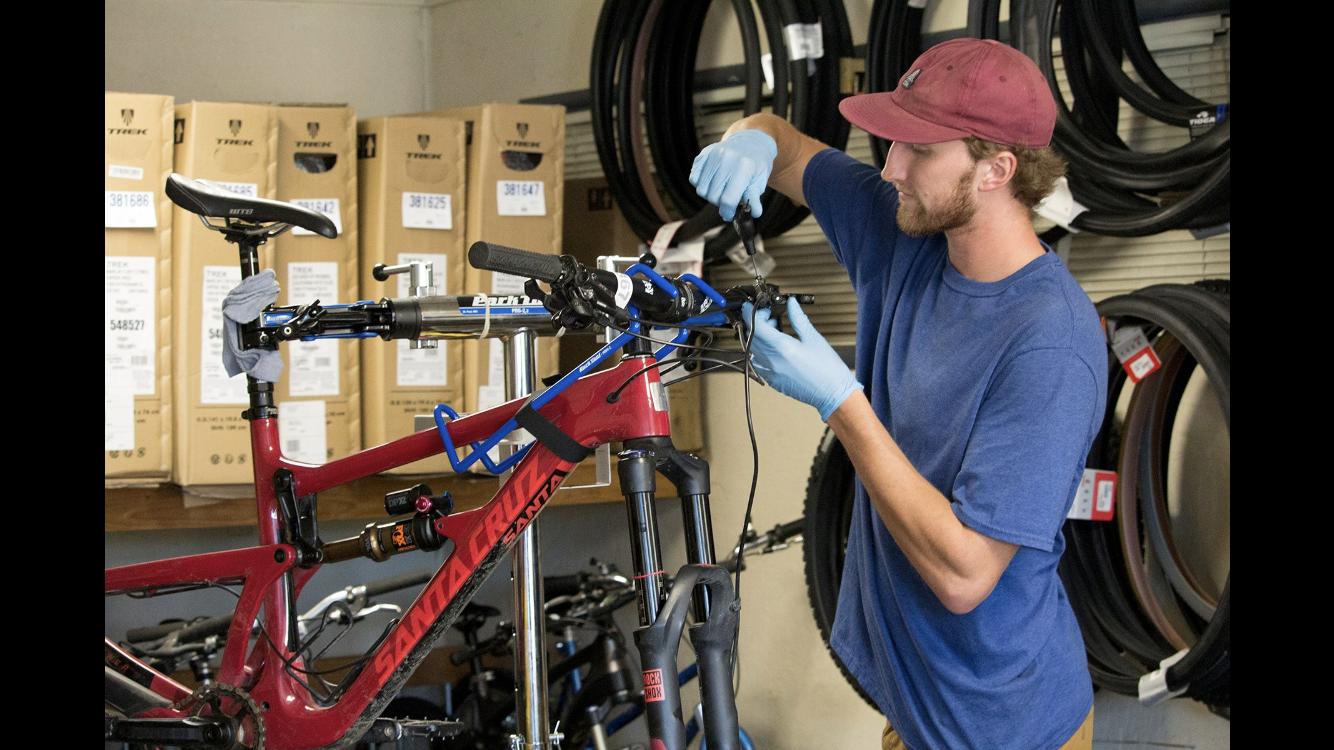 A bicycle technician wearing gloves is working on a red mountain bike in a shop. He is adjusting the handlebars while surrounded by boxes of bikes and hanging tires in the background.