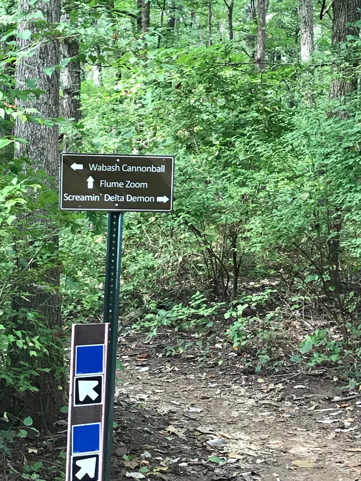 Signpost at a forest trail indicating directions to "Wabash Cannonball," "Flume Zoom," and "Screamin' Delta Demon," surrounded by dense green foliage. Percy Warner Mountain Bike Trails mountain bike trail.