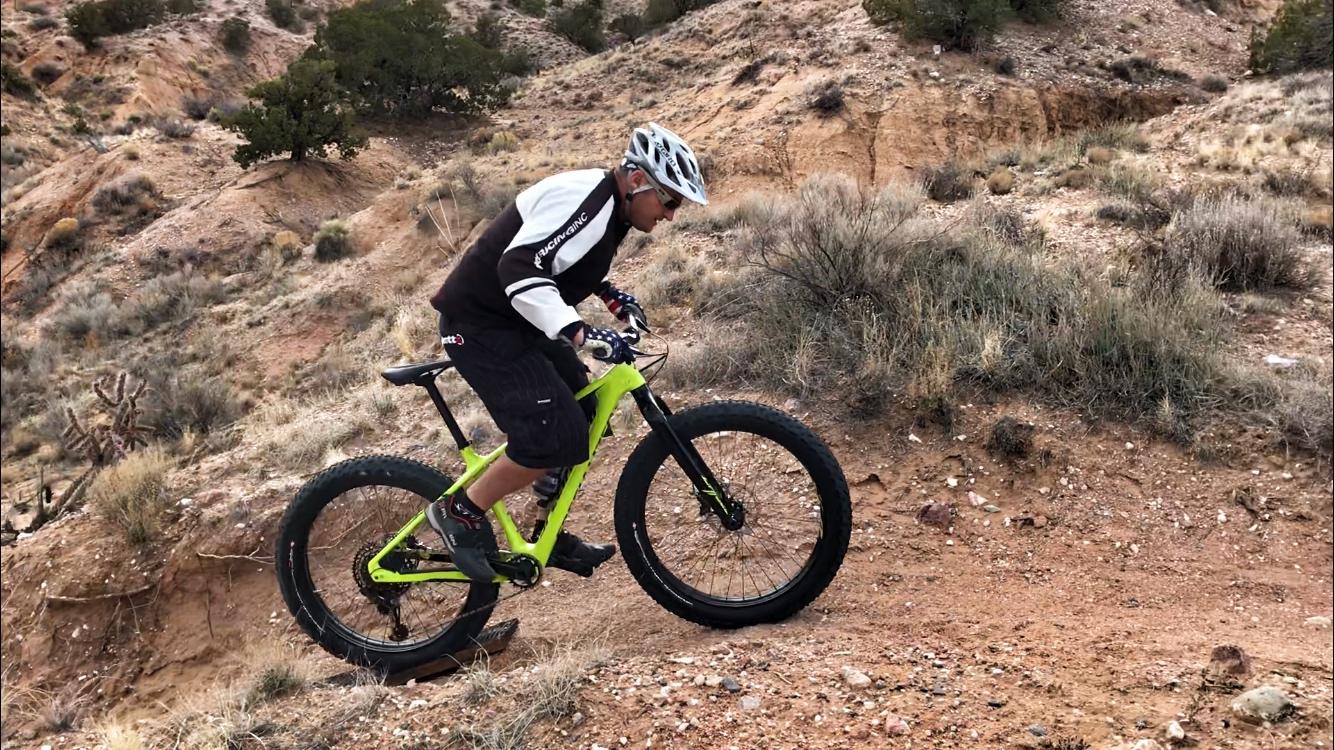 A person riding a bright green fat bike on a rocky, uneven dirt trail in a desert landscape. The cyclist is wearing a helmet and protective gear, and is focused on navigating the terrain. Surrounding vegetation includes small shrubs and cacti, with dry, hilly terrain in the background. Mariposa Fat Bike Trails mountain bike trail.
