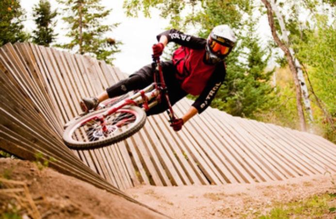 Specialized Stumpjumper FSR comp carbon 650B: A mountain biker in protective gear performs a sharp turn on a wooden ramp in a forested area. The rider, dressed in a red and black outfit, leans into the turn with one foot extended and the bike's tires close to the ground. Surrounding trees indicate a natural outdoor setting, highlighting the thrill of the sport.