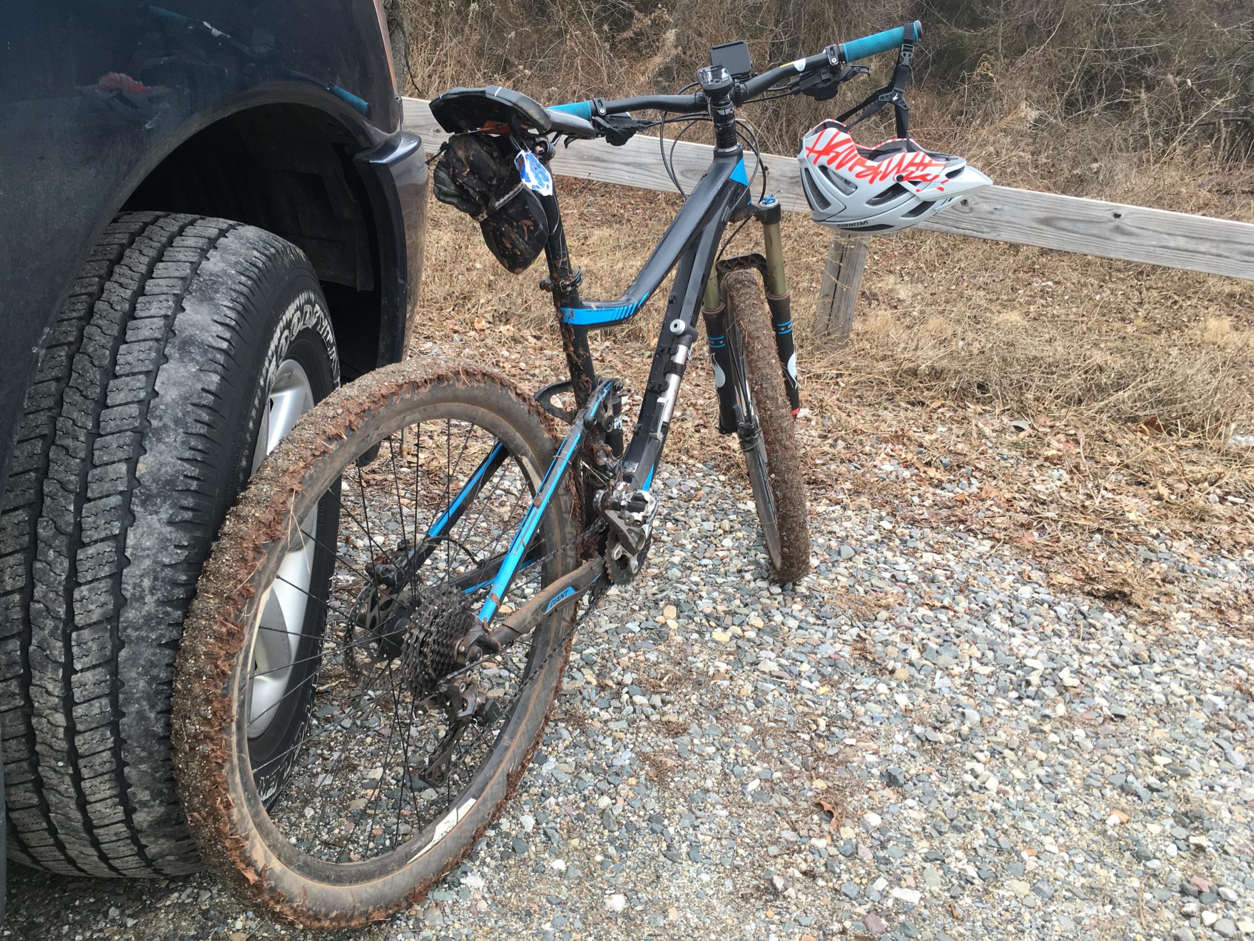 A mountain bike resting against a vehicle tire, with muddy tires and a helmet hanging from the handlebars. The background features gravel and dried vegetation, indicating an outdoor setting. Six Mile Run mountain bike trail.