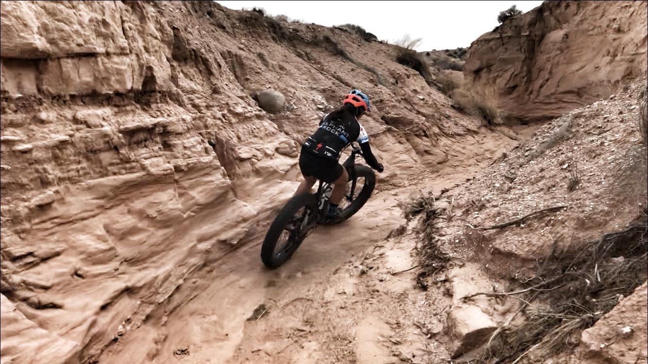 A person riding a fat bike through a narrow dirt trail in a canyon, surrounded by rocky terrain and sandy ground. The cyclist is wearing a helmet and athletic clothing, showcasing an action-packed moment in mountain biking. Mariposa Fat Bike Trails mountain bike trail.
