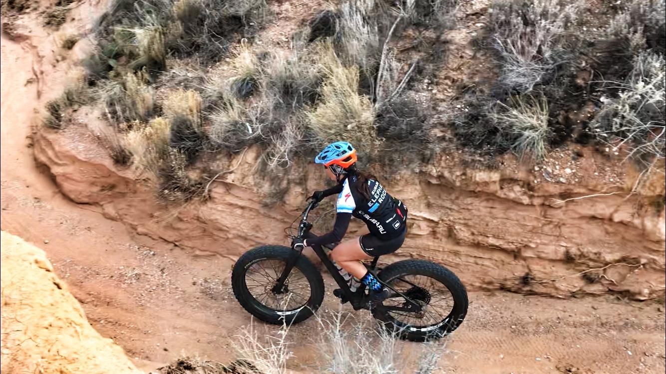 A cyclist riding a fat bike on a rocky trail, surrounded by dry vegetation and sandy terrain. The cyclist is wearing a helmet and a cycling jersey while navigating a curve in the path. Mariposa Fat Bike Trails mountain bike trail.